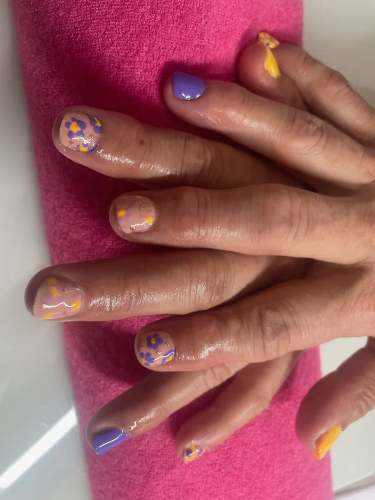 Manucure aux tons violet, jaune et nude, avec motifs floraux peints à la main, sur fond rose.